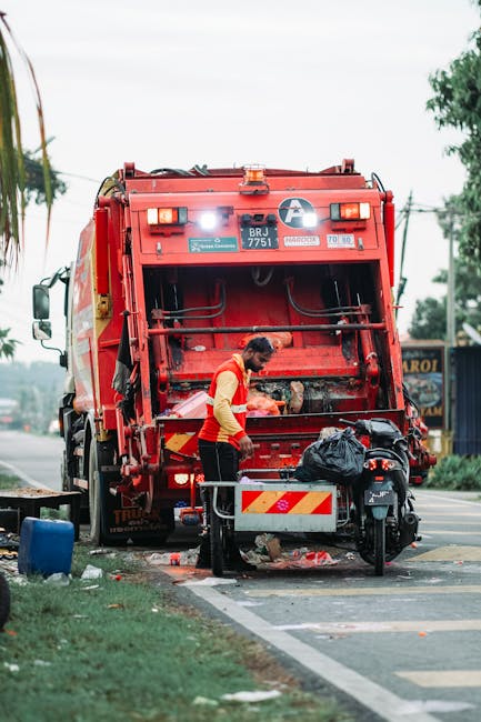 A worker wearing a red and yellow high-visibility vest is standing next to a large red rubbish collection truck parked on the side of a street. The truck's rear compactor mechanism is open, revealing 