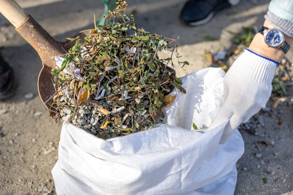 A narrow urban alleyway filled with a large accumulation of mixed waste and rubbish. In the foreground, a black wheeled bin made of durable plastic or metal is partially covered by a grey fabric bag, 