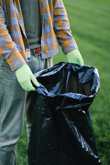 Three large black rubbish bags made of thick plastic material are stacked on a pavement beside a metal fence with vertical bars and a dark wooden or metal structure in the background. The bags appear